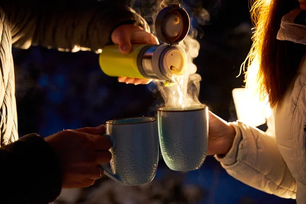 A couple warming up with hot tea in the evening winter woods in the light of car headlights.