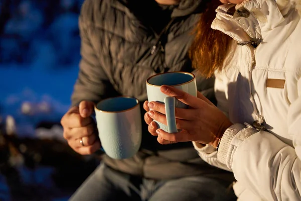 A couple warming up with hot tea in the evening winter woods in the light of car headlights.