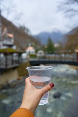 Borjomi şehrinin arka planında bir dağ pınarından bir bardak saf su tutan birinci şahıs manzarası.