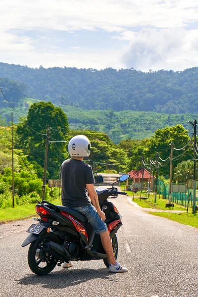 stock image A tourist guy on a motorbike stands in the middle of the road overlooking the jungle. Tropical exotic island in Asia. Langkawi, Malaysia - 06.20.2020