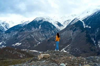Sarı ceketli ve kot pantolonlu bir kız karla kaplı dağların soğuk dağ manzarasının tadını çıkarıyor..