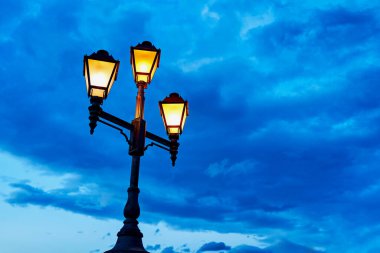 The lamppost of the city lights of the old district of the city. The warm light of the lantern against the blue cold sky.