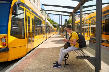 Sarı tişörtlü bir adam halka açık bir durakta tramvayını bekliyor. Etrafında sarı tramvaylar var..