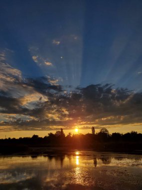 Nehir kenarında gün batımı manzarası, gökyüzünde güneş ışınları....