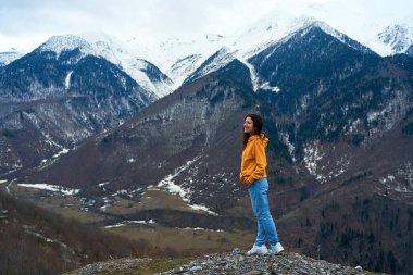 A girl in a yellow jacket and blue jeans enjoys the cold mountain scenery of snow-covered mountains.