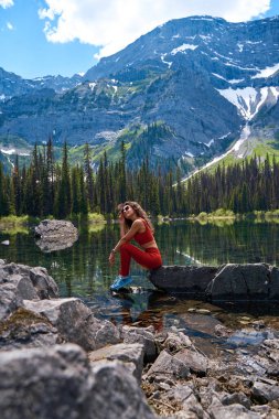 Spor takım elbiseli güzel, ince kadın, sakin, berrak gölde büyük bir taşın üzerinde poz veriyor. Yazın karlı yüksek dağların yansıması. Kananaskis Alberta 'da yürüyüş yaparken su kenarında dinlenen genç bir kız.