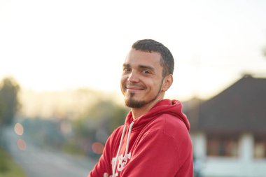 Sakallı, gülümseyen genç bir adamın portresi. Kırmızı kazaklı bir adam gün batımına karşı duruyor.