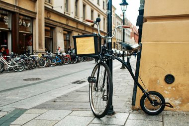 Alışılmadık bir asimetrik bisiklet Avrupa sokaklarında bir kaldırıma zincirlenmiş duruyor. Büyük ve küçük tekerli bir bisiklet.