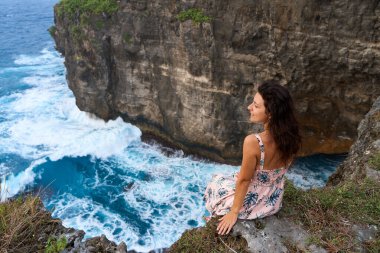 Pembe elbiseli güzel bir kadın Nusa Penida adasındaki okyanusun üzerindeki bir uçurumda oturuyor. Şeytan 'ın Billabong' u inanılmaz derecede harika bir göl.