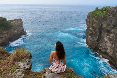 Pembe elbiseli güzel bir kadın Nusa Penida adasındaki okyanusun üzerindeki bir uçurumda oturuyor. Şeytan 'ın Billabong' u inanılmaz derecede harika bir göl.