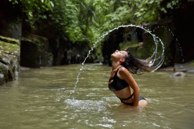Mayo giymiş genç ve narin bir kadın popüler Bali adasındaki bir dağ nehrinin kenarında poz veriyor.