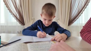 Evde elinde kalemle kitap yazan bir çocuk annesiyle ödev yapıyor.