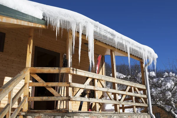 带雪冠的木制房屋 屋顶上的大冰柱 阳台上的滑雪设备 — 图库照片