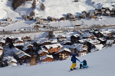 İki snowboardcu yüksek kış dağlarında karlı kayak yamaçlarında ve arka planda güneşli bir köyde. İtalyan Alplerinde akşam yemeği. Levigno, Lombardiya bölgesi, İtalya, Avrupa.