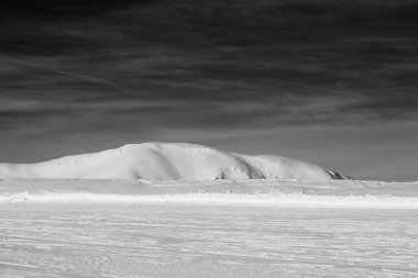 Snowy ski slope at high winter mountains and sunlit cloudy sky. Italian Alps. Livigno, region of Lombardy, Italy, Europe. Selective focus on background. Black and white toned landscape.