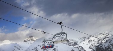 Sunlit chair lift, snowy ski slope, high winter mountains and dark cloudy sky. Ski area Mottolino Fun Mountain, Italian Alps. Livigno, Lombardy, Italy, Europe. Panoramic view.