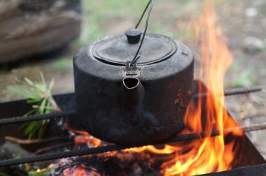 Sooty old teakettle on bonfire with smoke at forest in sunny summer day. Selective focus.