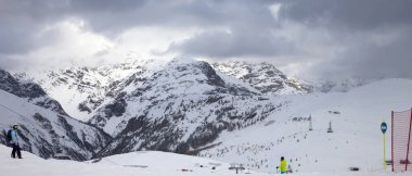 Snowy ski slope with skier and snowboarder in high mountains and cloudy sunlit sky at winter gray day. Ski area Mottolino Fun Mountain, Italian Alps. Livigno, Lombardy, Italy, Europe. Panoramic view.