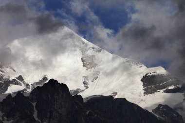 High snowy mountains with glacier, rocks and blue sky with dark clouds before storm. Caucasus Mountains. Georgia, region Svanetia, Mount Tetnuldi.