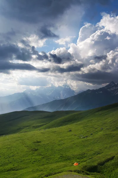 Orange tourist tent on green meadow in high mountains and sunlit cloudy sky. Caucasus Mountains. Georgia, region Svanetia in summer. 
