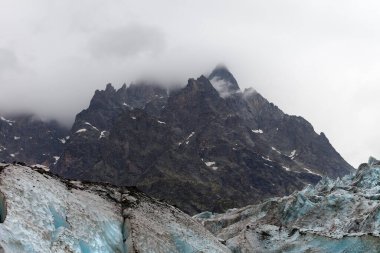 High rocky mountains in fog and glacier with deep danger crevasses covered with snow and ground at gray day. Caucasus Mountains. Georgia, region Svanetia.