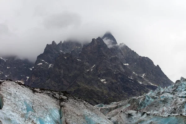 High rocky mountains in fog and glacier with deep danger crevasses covered with snow and ground at gray day. Caucasus Mountains. Georgia, region Svanetia.