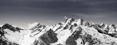 Panorama of snow-capped mountain peaks and sky with clouds at sun windy day. Caucasus Mountains in winter, region Dombay. Black and white toned landscape.