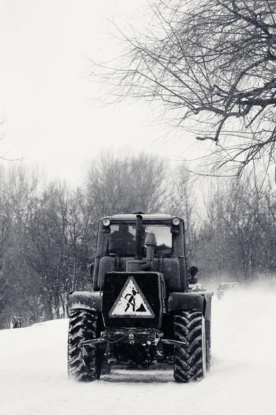Old tractor clears the snow-covered road from snow blockages. Snowy country road through forest in bad weather after snowfall. Black and white toned image.