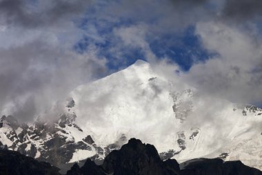 High snowy mountains with glacier, rocks and blue sky with dark fog before storm. Caucasus Mountains. Georgia, region Svanetia, Mount Tetnuldi.