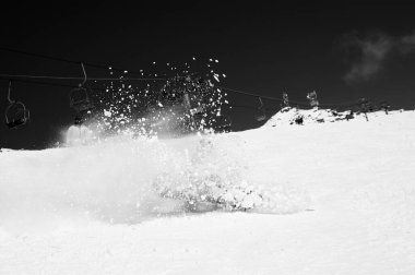 Snowboarder fall down with snow splashes on snowy ski slope and chair lift at background. Caucasus Mountains in sunny winter day, region Dombay. Black and white toned image. High contrast