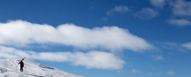 Blue sky and skier with skis on his shoulder go up to top of mountain. Caucasus Mountains in winter, Georgia, region Gudauri, Mount Kudebi. Panoramic view
