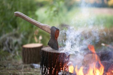 Ağaç kütüğünde balta ve yaz ormanında gece dumanla kamp ateşi.