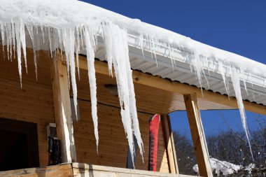 Wooden house with snow cornice and big icicles on roof and ski equipment on balcony at sunny winter day clipart