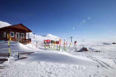 Yüksek karlı dağlar, ahşap kurtarma evi, kar yığınlarında uyarı işaretleri, arka planda teleferik ve güneş ışığıyla mavi gökyüzü. Güneşli kış gününde Kafkas Dağları. Gürcistan, bölge Gudauri.