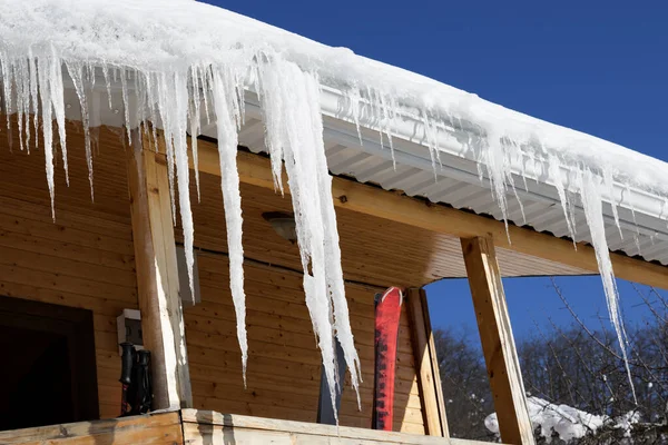 日当たりの良い冬の日にバルコニーに屋根やスキー用具に雪のコーニスと大きなつららを持つ木造の家 — ストック写真