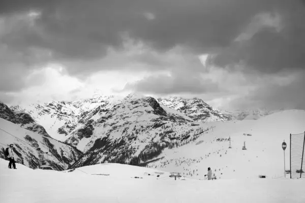Black and white snowy ski slope with skier and snowboarder in high mountains and cloudy sunlit sky at winter gray day. Ski area Mottolino Fun Mountain, Italian Alps. Livigno, Lombardy, Italy, Europe.