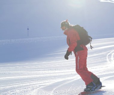 Kırmızılı snowboardcu, kış akşamı güneşli bir kış akşamında bakım makinesinin hazırladığı karlı kayak pistine iniyor.