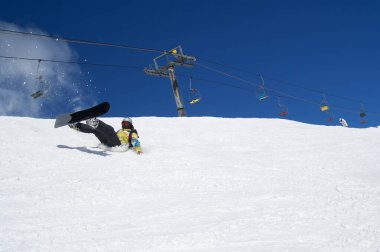 Snowboardcu, arka planda karlı kayak pistine kar damlaları ve sandalye kaldırmasıyla yere düşüyor. Güneşli kış gününde Kafkasya Dağları Dombay bölgesi.