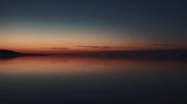 Sakin denizin üzerinde huzurlu bir günbatımı. Canlı turuncu ve mavi eğimli gökyüzü durgun suyu yansıtır. Alacakaranlıkta çekilen minimalist deniz manzarası, arka plan veya meditasyon görüntüleri için ideal.