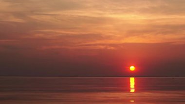 Sakin denizin üzerinde huzurlu bir günbatımı. Canlı turuncu ve mavi eğimli gökyüzü durgun suyu yansıtır. Alacakaranlıkta çekilen minimalist deniz manzarası, arka plan veya meditasyon görüntüleri için ideal.