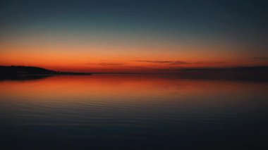 Sakin denizin üzerinde huzurlu bir günbatımı. Canlı turuncu ve mavi eğimli gökyüzü durgun suyu yansıtır. Alacakaranlıkta çekilen minimalist deniz manzarası, arka plan veya meditasyon görüntüleri için ideal.