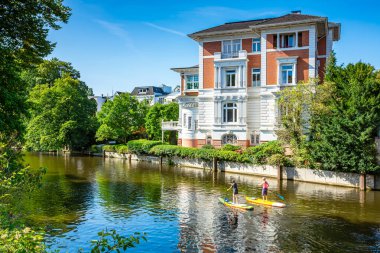 Nehirdeki ev, güneşli bir yaz gününde nehirde sörf yapan insanlar. Hamburg, Almanya 'da nehir kenarındaki güzel evler.