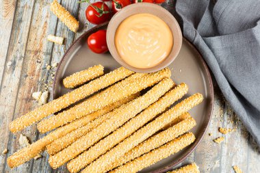 Kahverengi seramik tabakta susamlı ekmek çubukları, peynir sosu, kiraz domatesleri ve ahşap masadaki gri kumaş peçetesi..