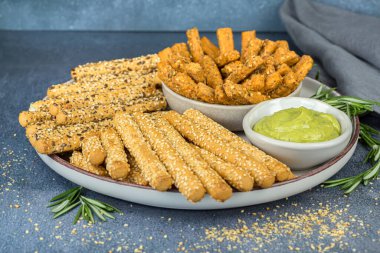 Ekmek çubukları kablosuz tohumlar, bitki sosu ve biberiye yaprakları seramik tabakta, kumaş peçete gri arka planda.