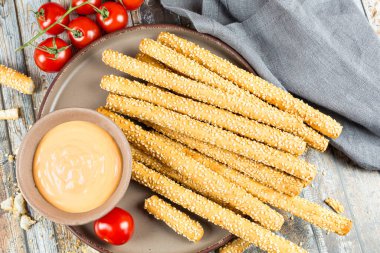 Kahverengi seramik tabakta susamlı ekmek çubukları, peynir sosu, kiraz domatesleri ve ahşap masadaki gri kumaş peçetesi..