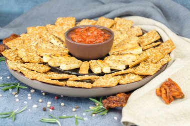 Kahverengi seramik tabakta domates soslu ekmek çubukları ve krakerler, gri arka planda kumaş peçeteler..