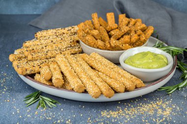 Ekmek çubukları kablosuz tohumlar, bitki sosu ve biberiye yaprakları seramik tabakta, kumaş peçete gri arka planda.