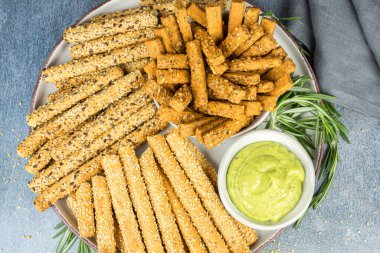 Ekmek çubukları, bitki sosu ve biberiye yaprakları seramik tabakta, kumaş peçete gri arka planda.