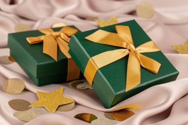 Pembe ipek arka planda sarı kurdeleli iki yeşil hediye kutusu. Pembe ipek arka planda yuvarlak konfeti ve sarı yıldızlar.