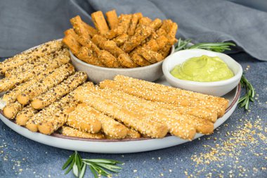 Ekmek çubukları kablosuz tohumlar, bitki sosu ve biberiye yaprakları seramik tabakta, kumaş peçete gri arka planda.